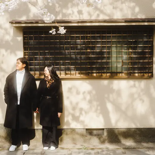Priam and Eden holding hands by a sunlit wall with cherry blossoms
